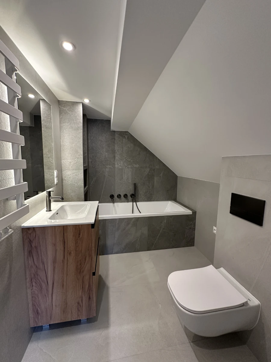 Mała nowoczesna łazienka z podświetlanym lustrem, widok z boku z wanną i szarymi płytkami