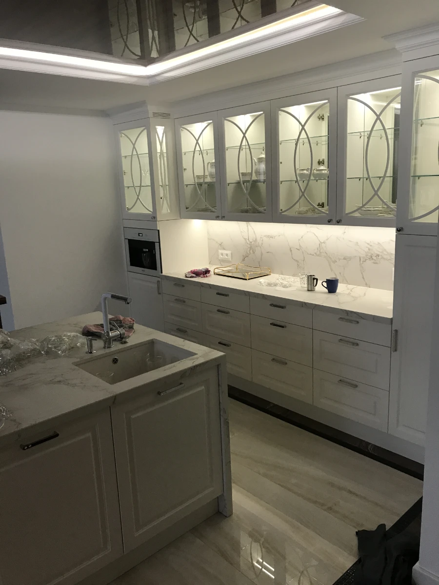 Fragment kuchni z białymi meblami i marmurowym blatem oraz ozdobnymi witrynami na porcelanę